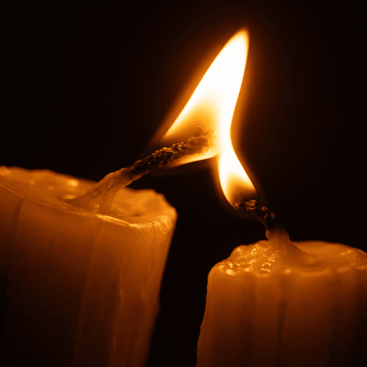 One candle flame leaning to kindle a second wick, the new flame catching in the dark
