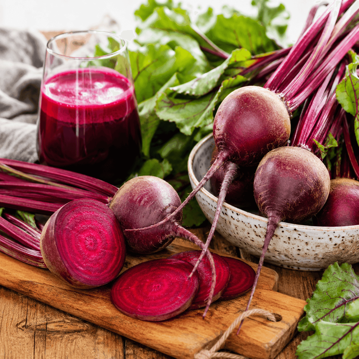 Whole and sliced red beets with their leafy greens in a speckled bowl beside a glass of fresh beet juice on a wooden board