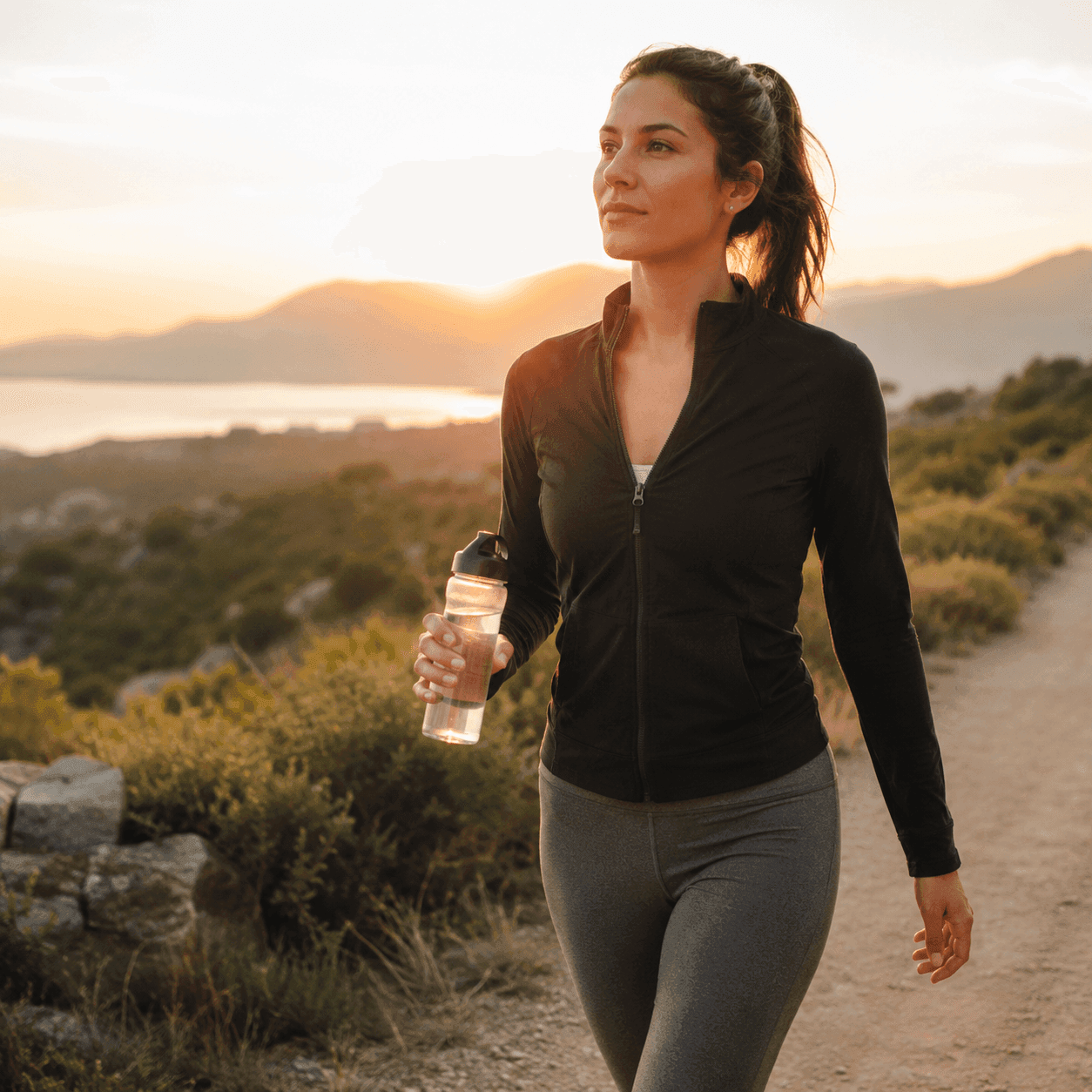 A woman walking on a coastal trail at sunset holding a water bottle